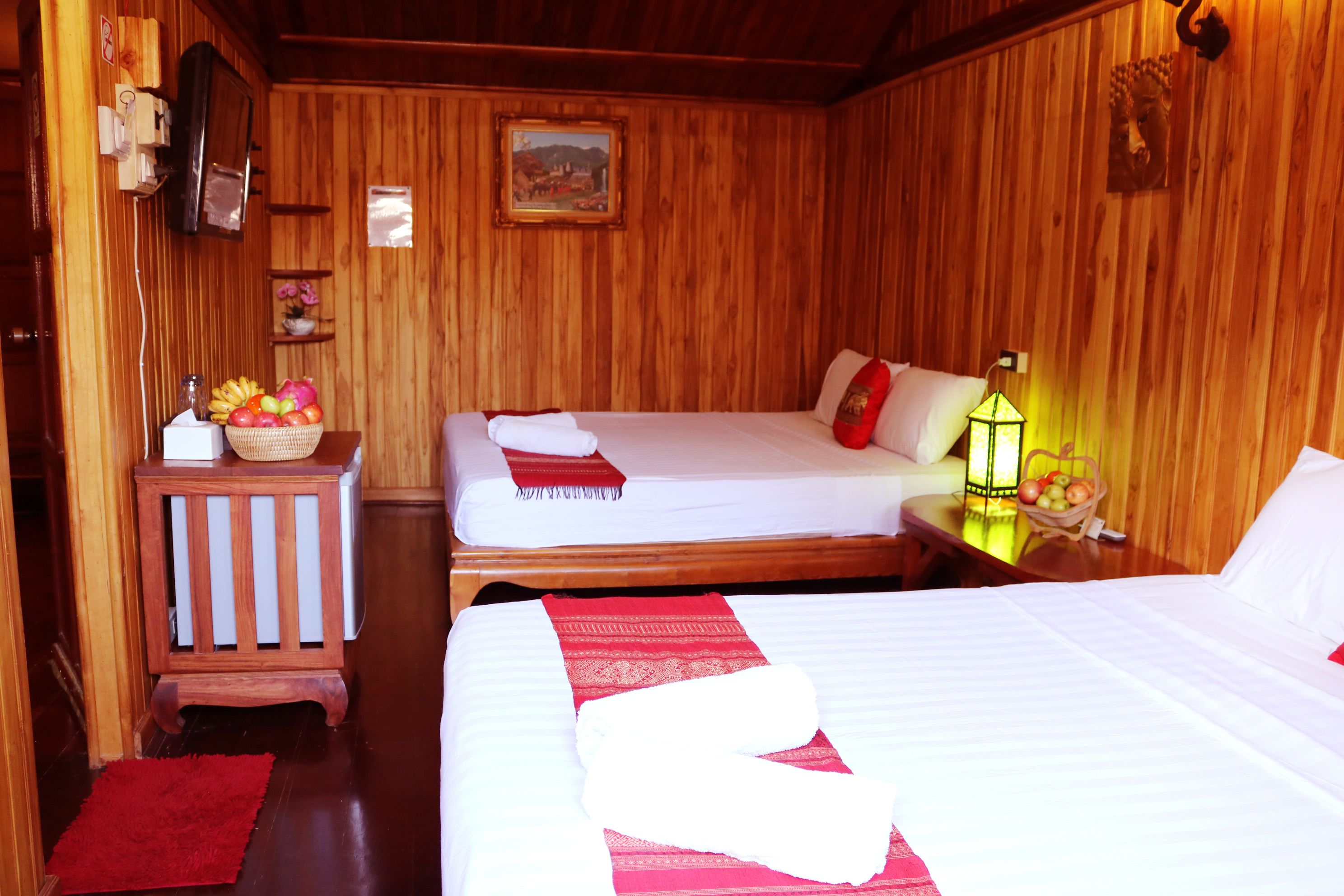 A wooden room with two beds, a small table with fruit, a red rug, and a wall-mounted TV.
