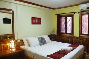 Una cama bien hecha con sábanas blancas y una manta estampada en rojo y verde, una mesita de noche de madera con un farol, un cuadro en la pared y una ventana con rejas decorativas de hierro.