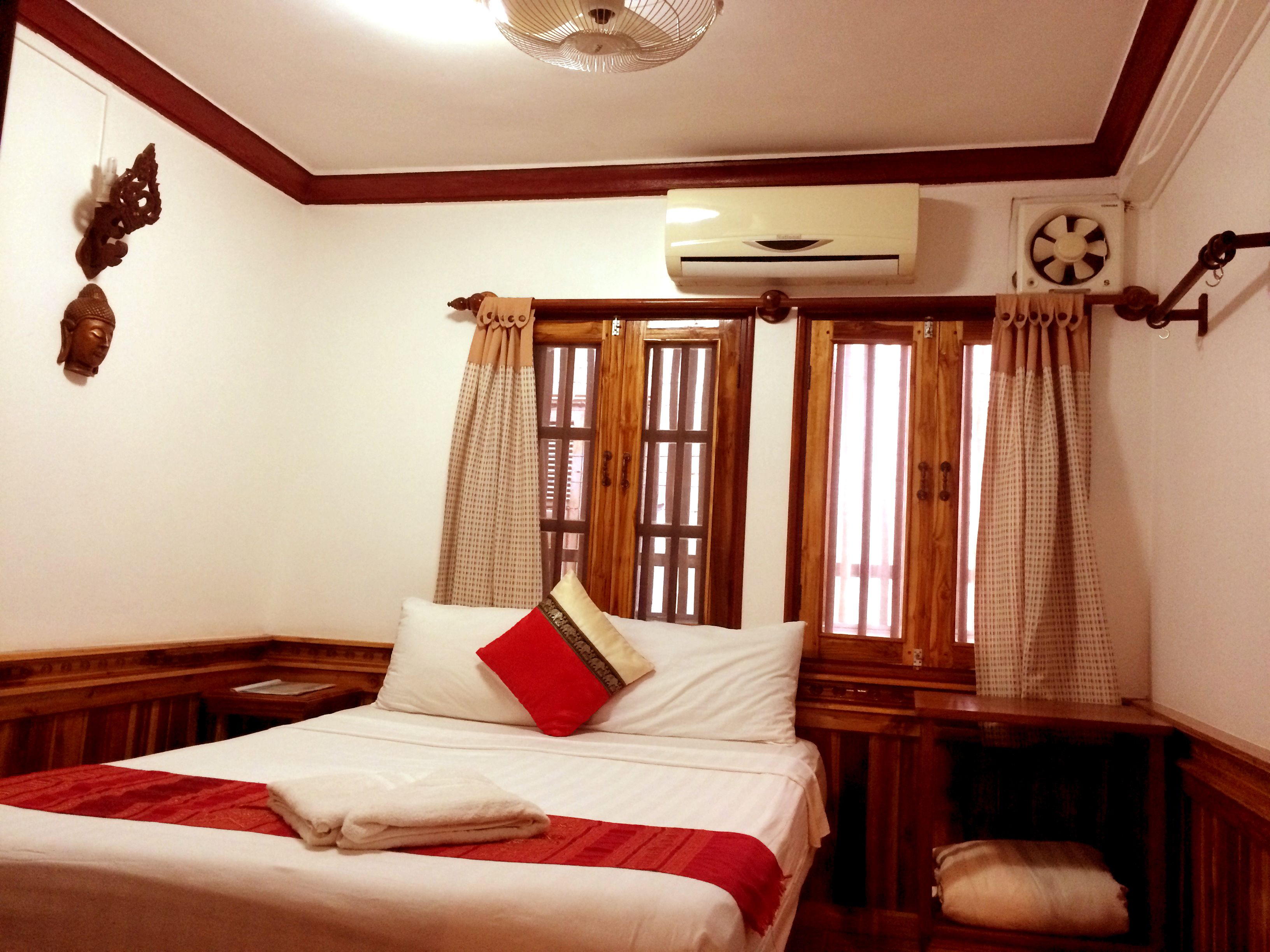 A neatly made bed with a red pillow, wooden headboard, and a white blanket. There is a wooden nightstand with a lamp, a wall-mounted air conditioner, and a window with curtains.