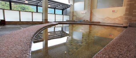 A spacious indoor swimming pool with clear water, surrounded by a tiled floor and walls, and large windows offering a view of greenery outside.