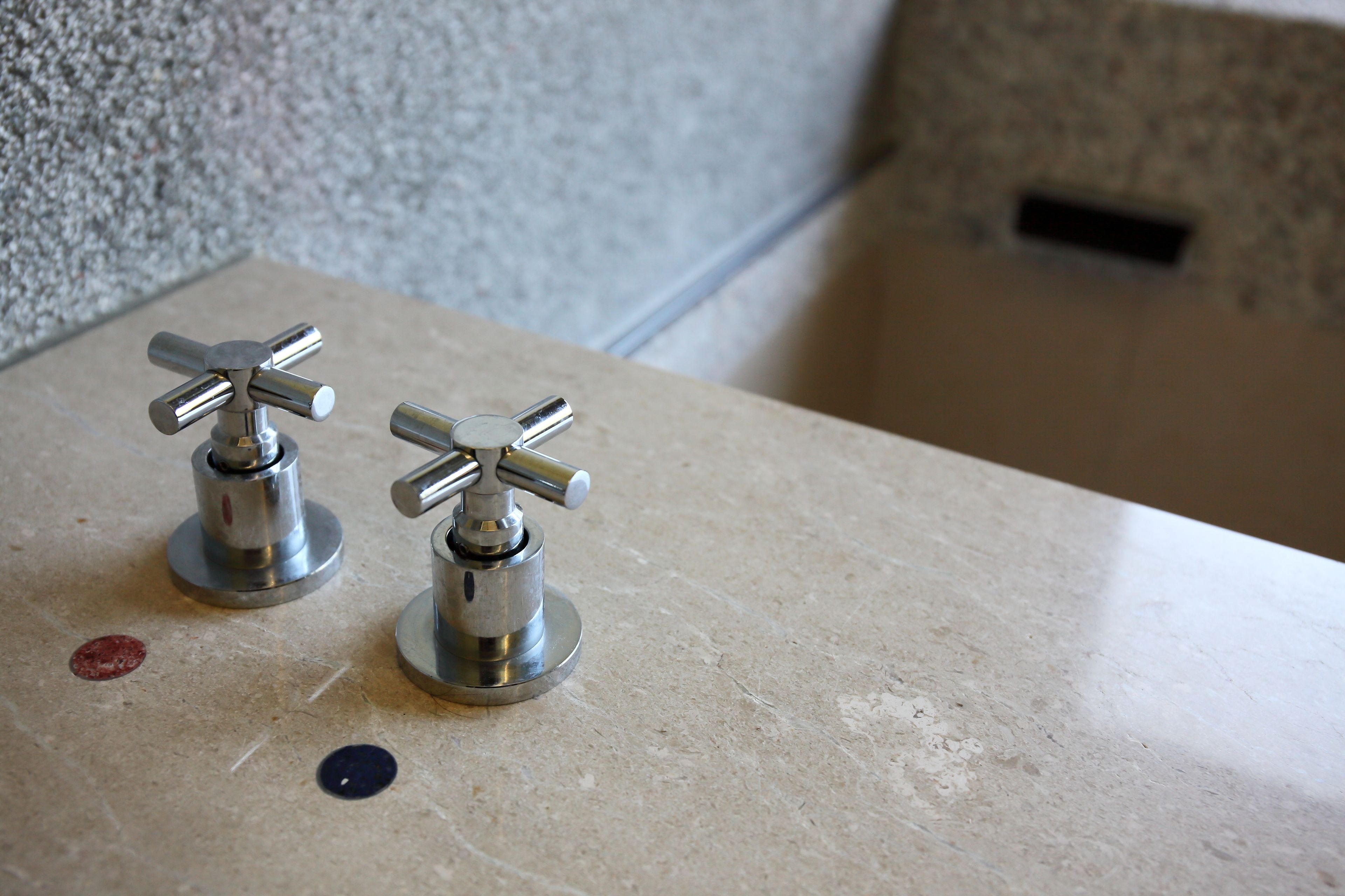 Twee metalen kraan(en) op een beige aanrecht met een gestructureerde muur op de achtergrond.