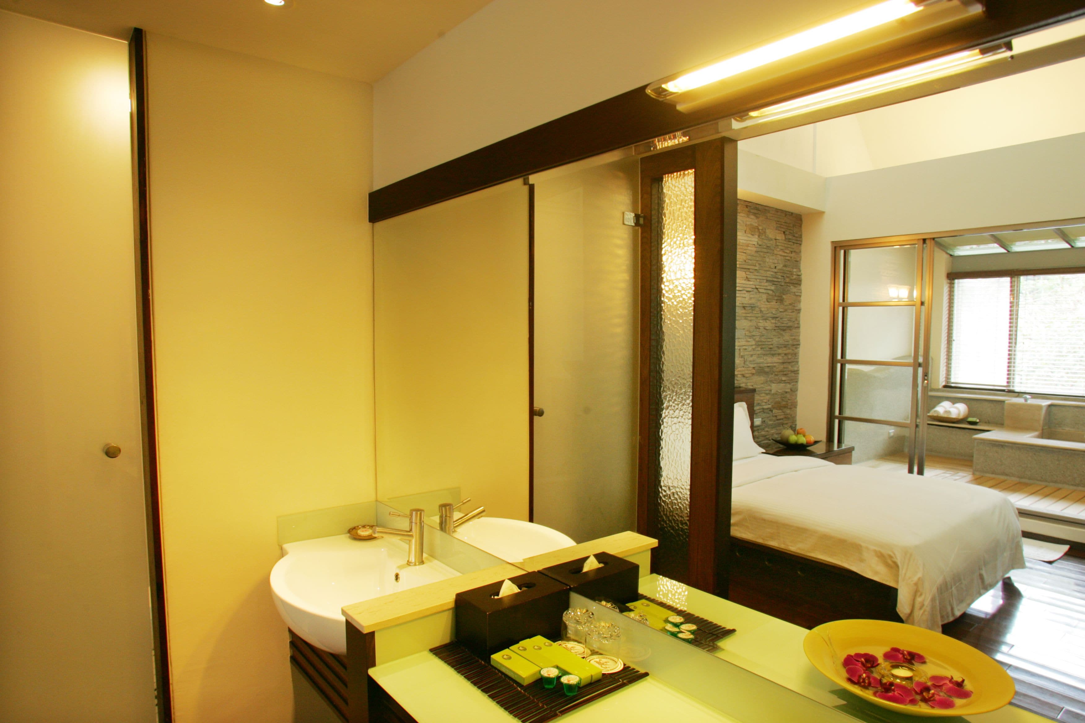 A hotel bathroom with a double sink, a large mirror, and a bed visible through an open door.