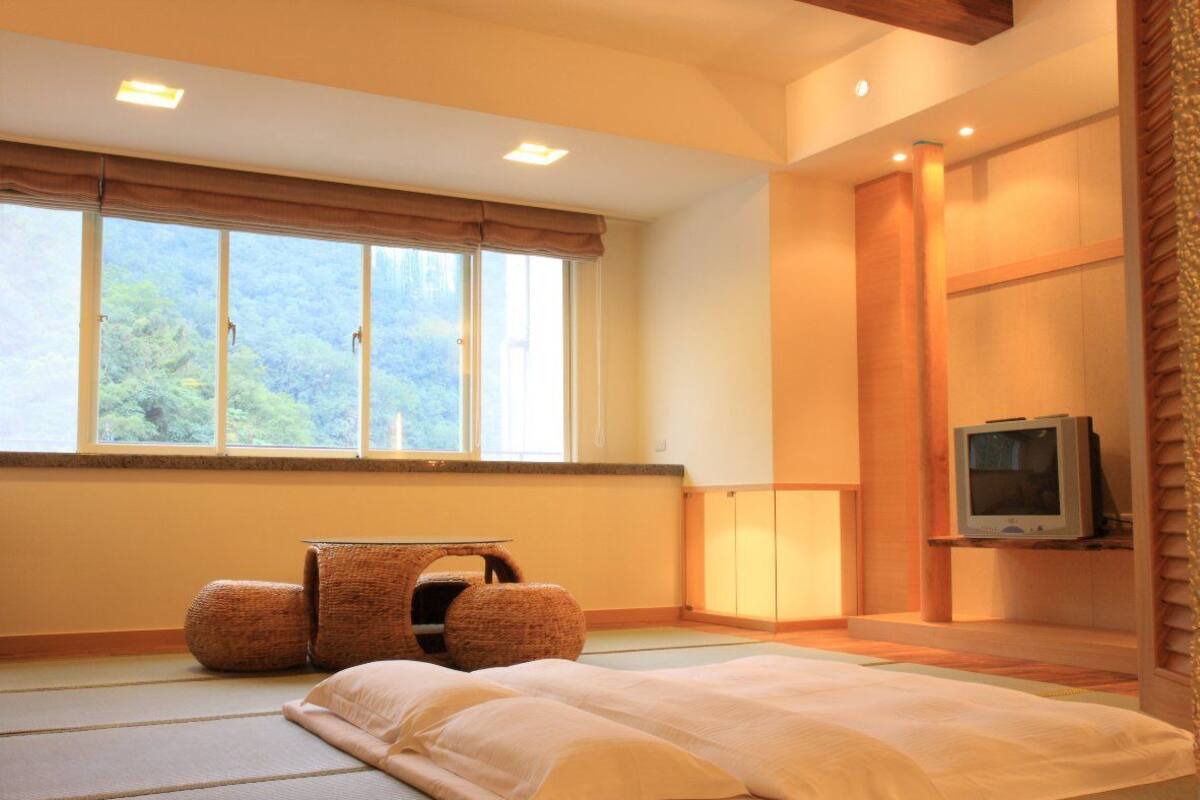 Une pièce avec des tatamis au sol, un futon servant de lit, une télévision et une vue sur la verdure environnante.