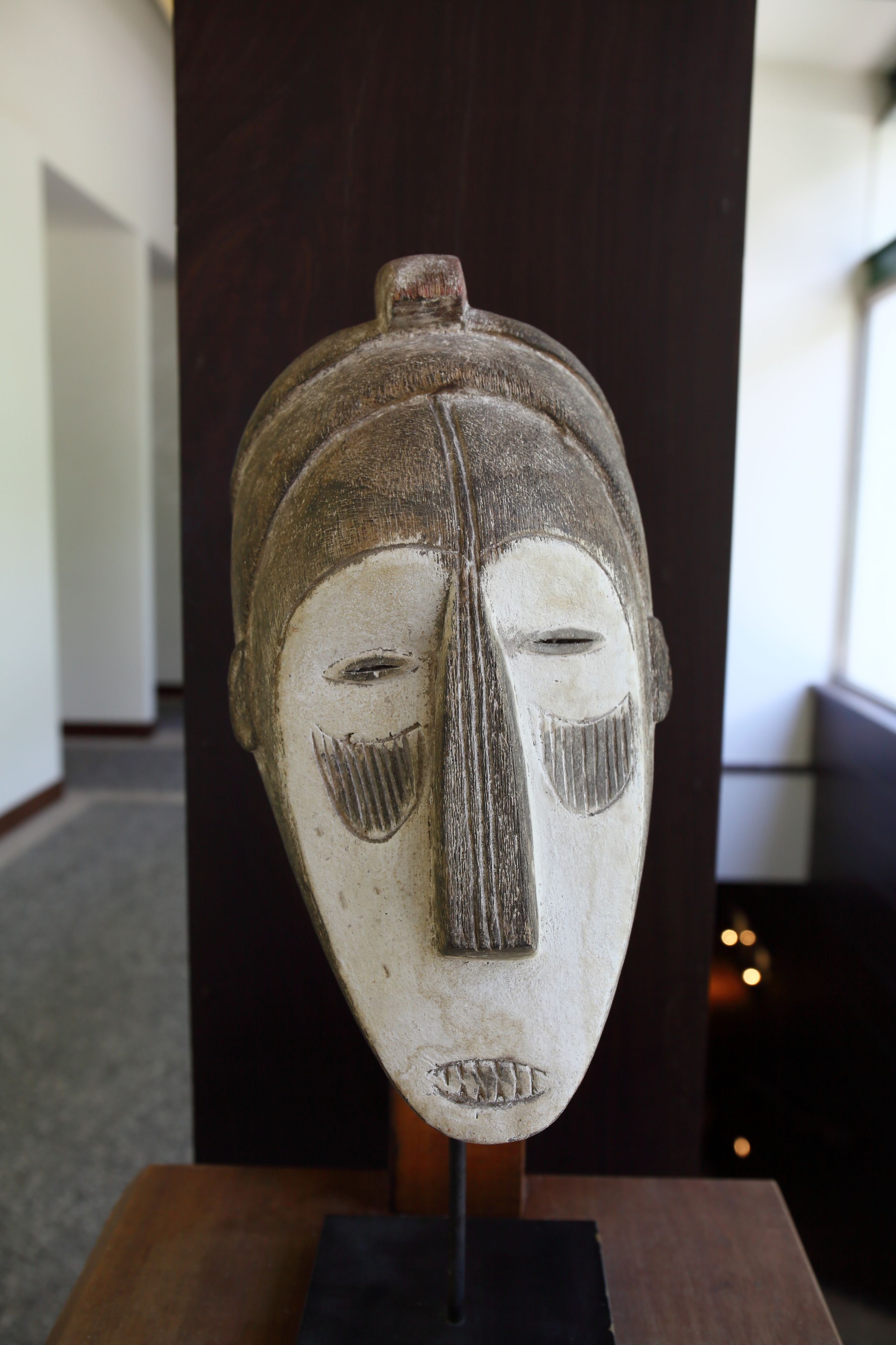 Una escultura de madera con rostro tallado, expuesta sobre un pedestal en una habitación con un pilar de madera oscura y suelo alfombrado.