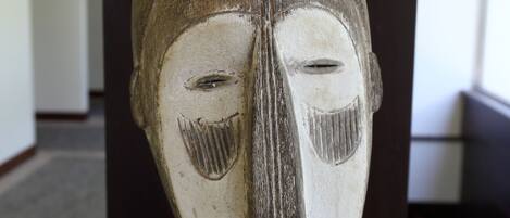 Una escultura de madera con rostro tallado, expuesta sobre un pedestal en una habitación con un pilar de madera oscura y suelo alfombrado.