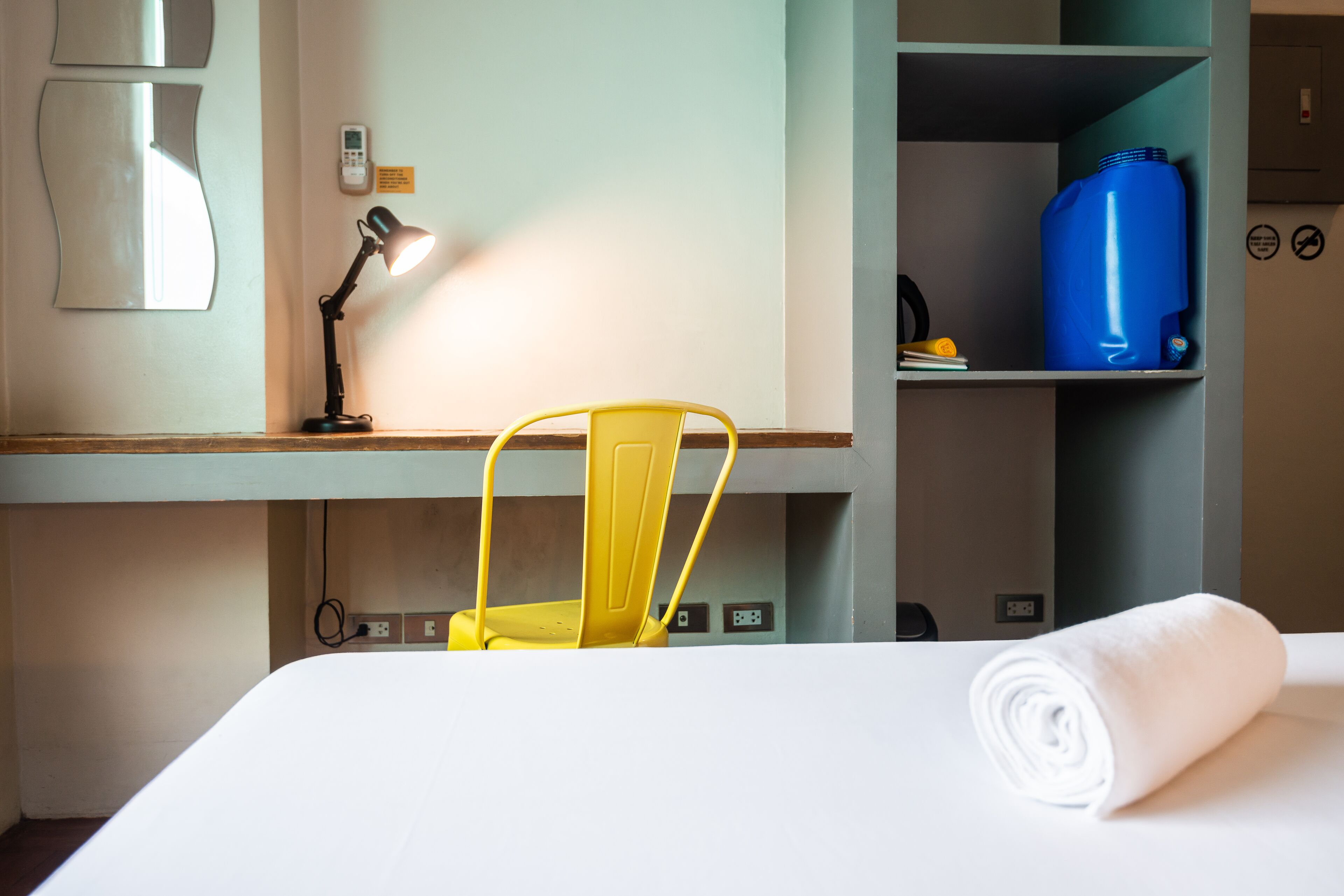 A hotel room with a bed, a desk with a lamp, a yellow chair, a blue water jug, and a folded towel.