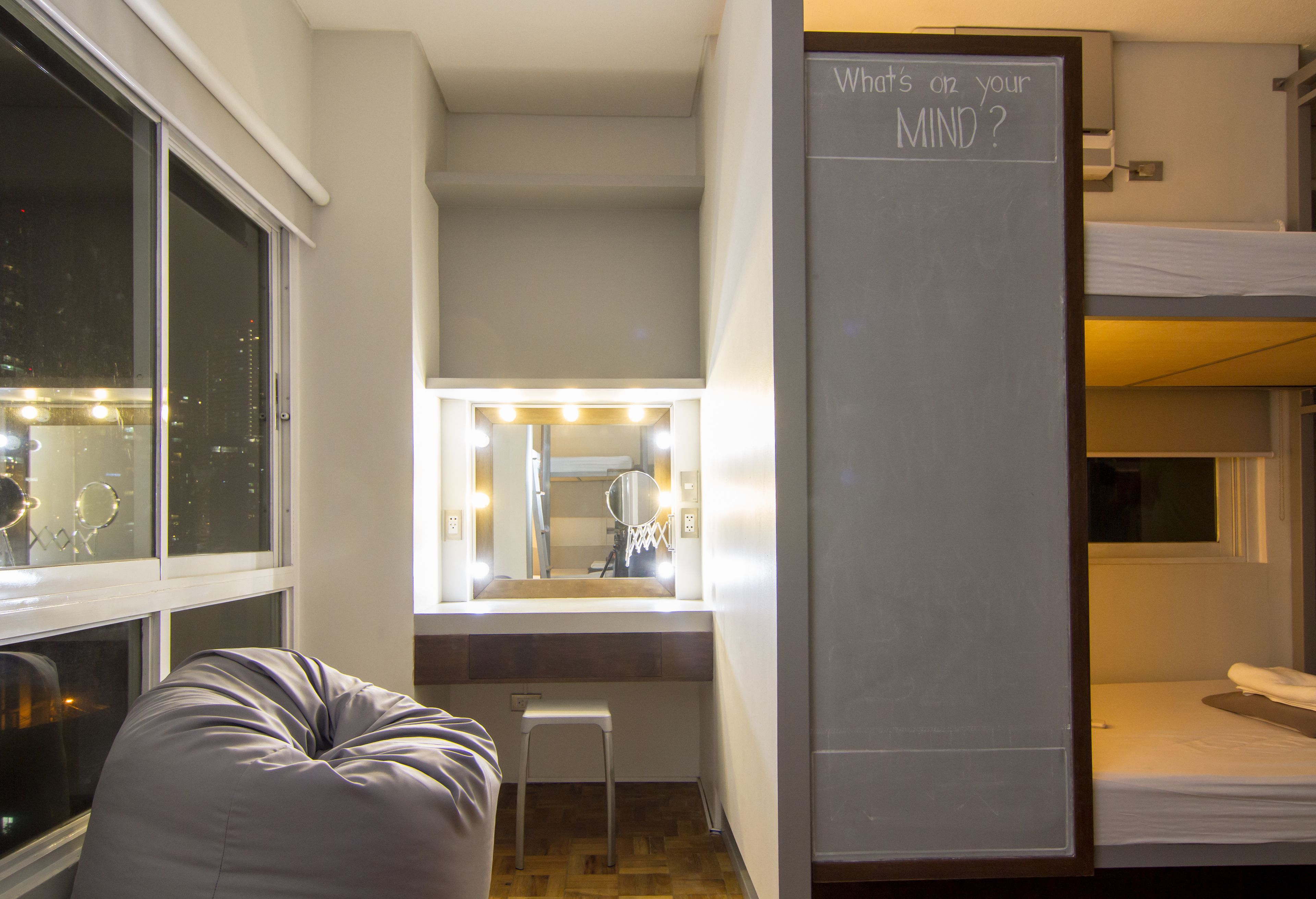 A hotel room with a vanity area, a mirror with lights, a bunk bed, and a window with a city view.