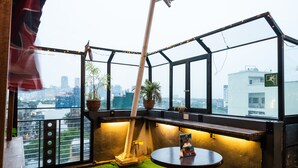 Une chambre située dans un immeuble de grande hauteur, offrant une vue sur la ville, avec une petite table sur laquelle repose un magazine et un sol recouvert d’un tapis d’herbe artificielle verte.