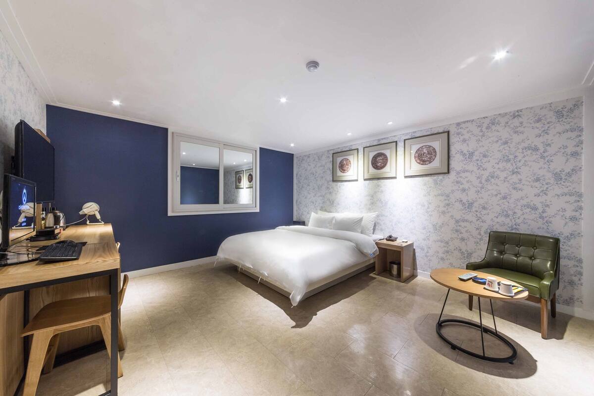 A bedroom with a bed, a desk with a computer, a chair, and a small table.