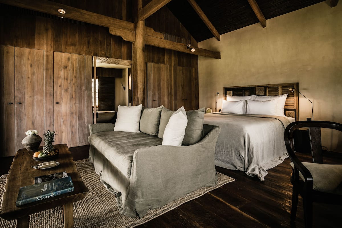 Un dormitorio acogedor con techo de madera, cama con ropa blanca, un sofá gris y una pequeña mesa con un florero y frutas.