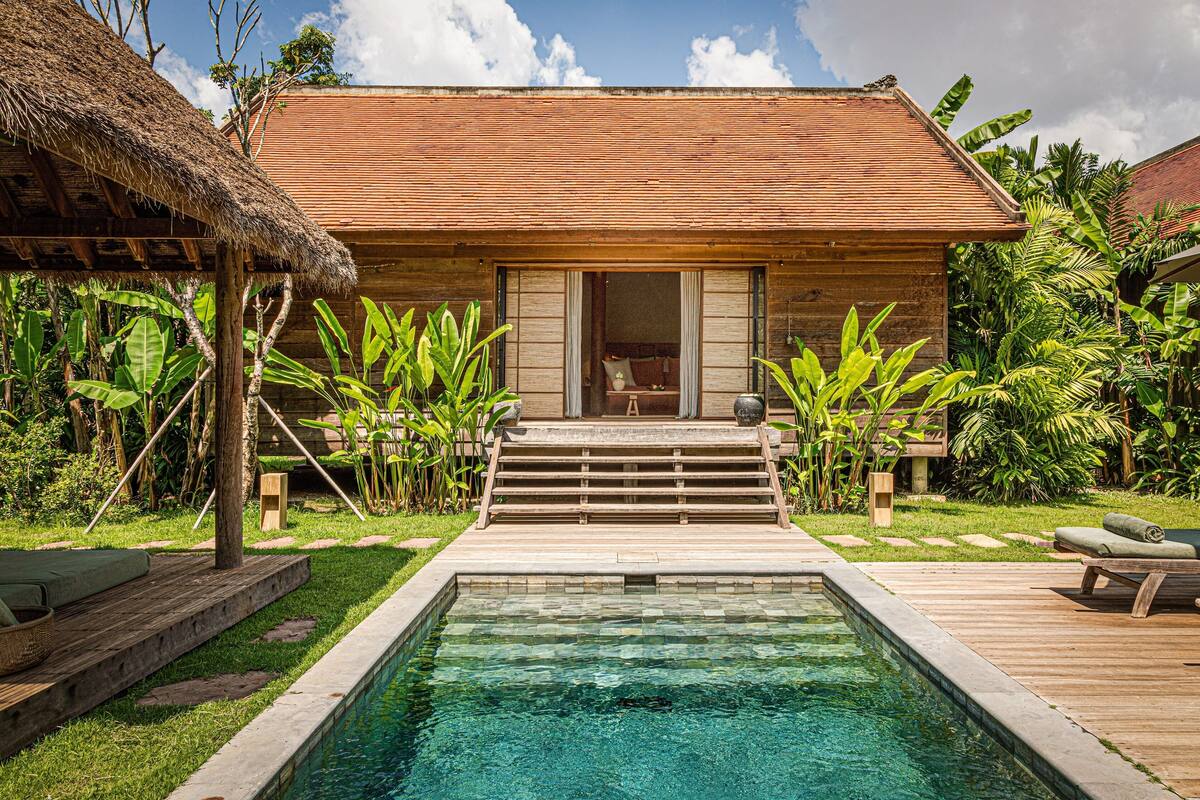 Una casa de madera con piscina, rodeada de exuberante vegetación y un tejado de paja.