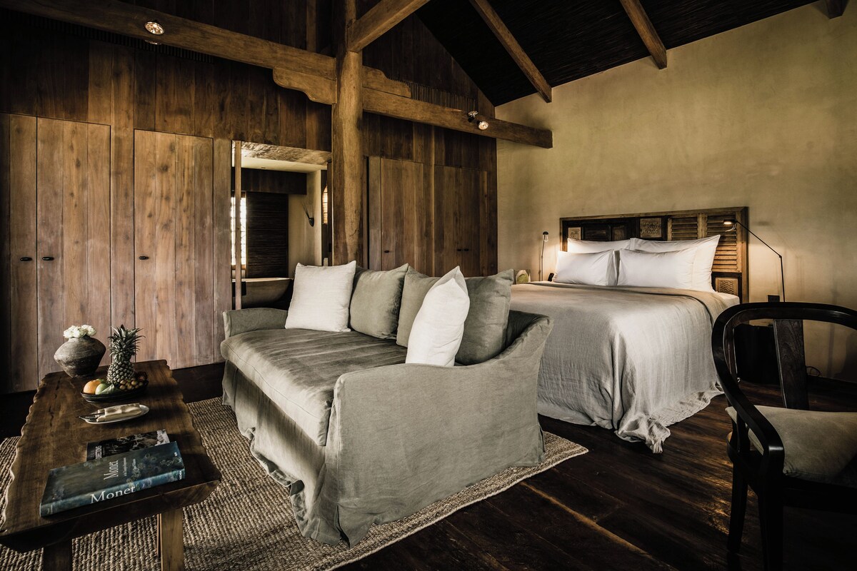 Een knus slaapkamer met een houten plafond, een bed, een bank en een kleine tafel met een vaas en fruit.