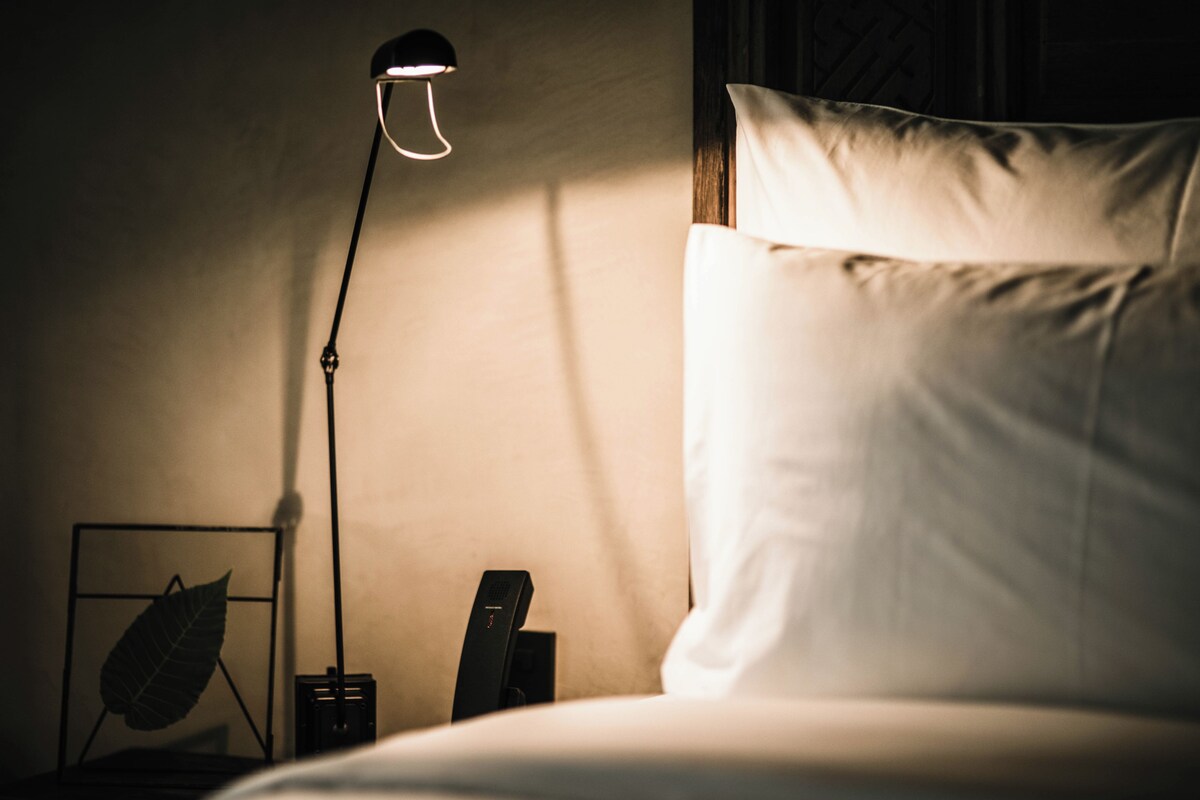 Una cama bien tendida con sábanas blancas, una mesita de noche con una lámpara y una decoración con hojas en un marco.