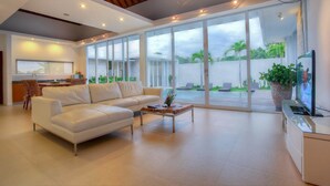 A spacious living room with a white leather sofa, a wooden coffee table, and a TV on a cabinet. Large sliding glass doors lead to an outdoor area with a pool and greenery. - Villa Umah Putih (Canggu)