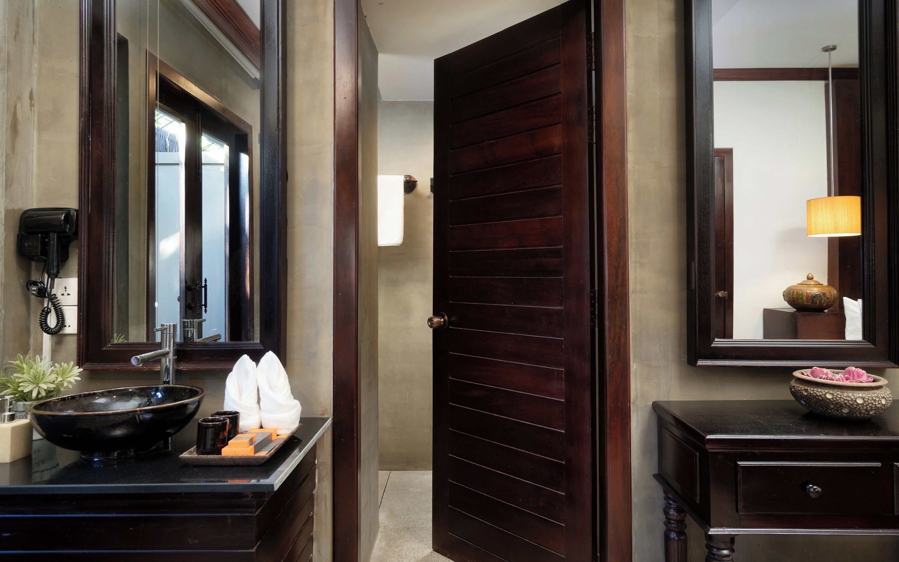 A hotel room with a dark wooden door, a bathroom with a large mirror, a sink with toiletries, and a small table with decorative items.