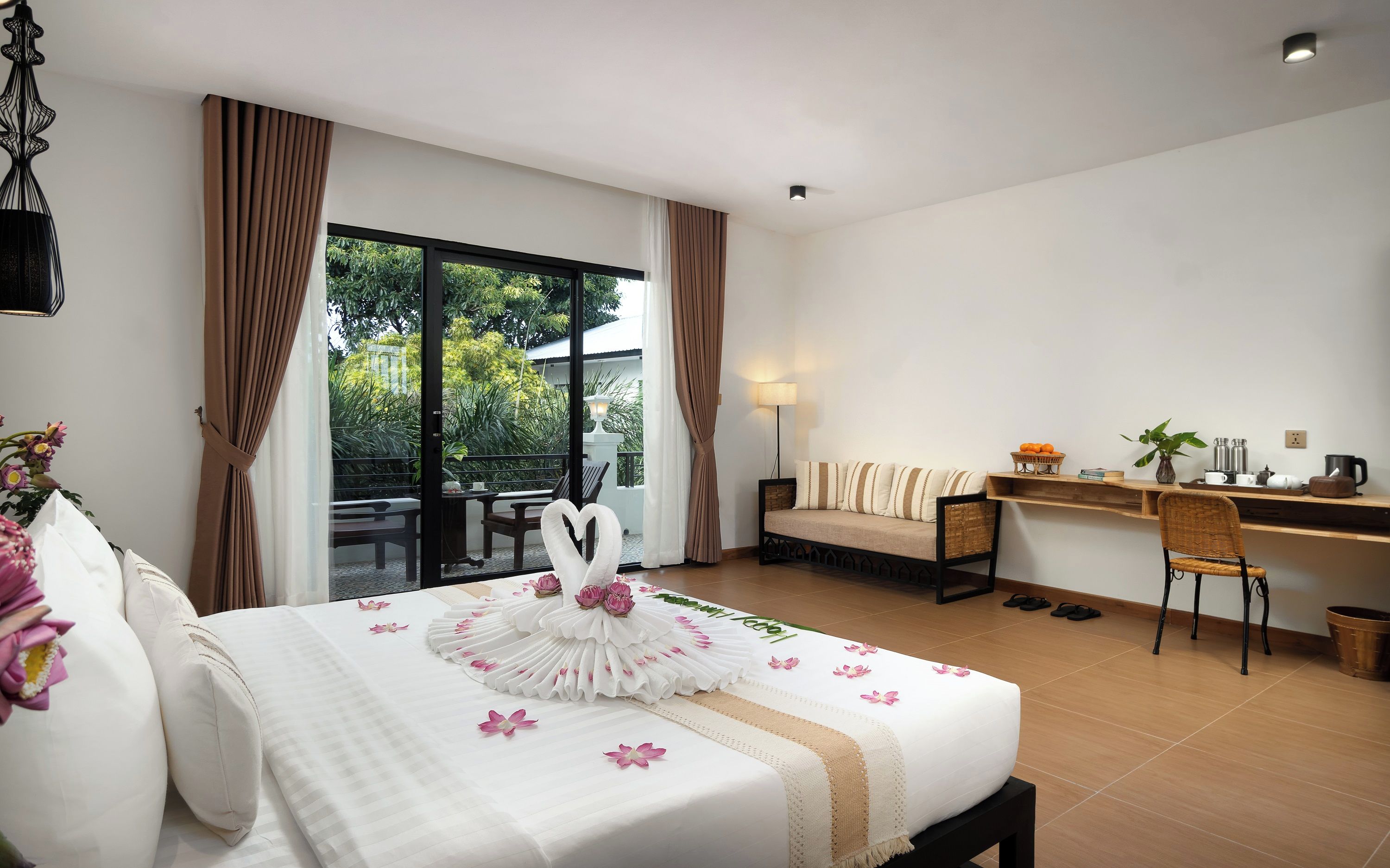 A hotel room with a bed, a desk, a chair, a balcony with a table and chairs, and a view of greenery outside.