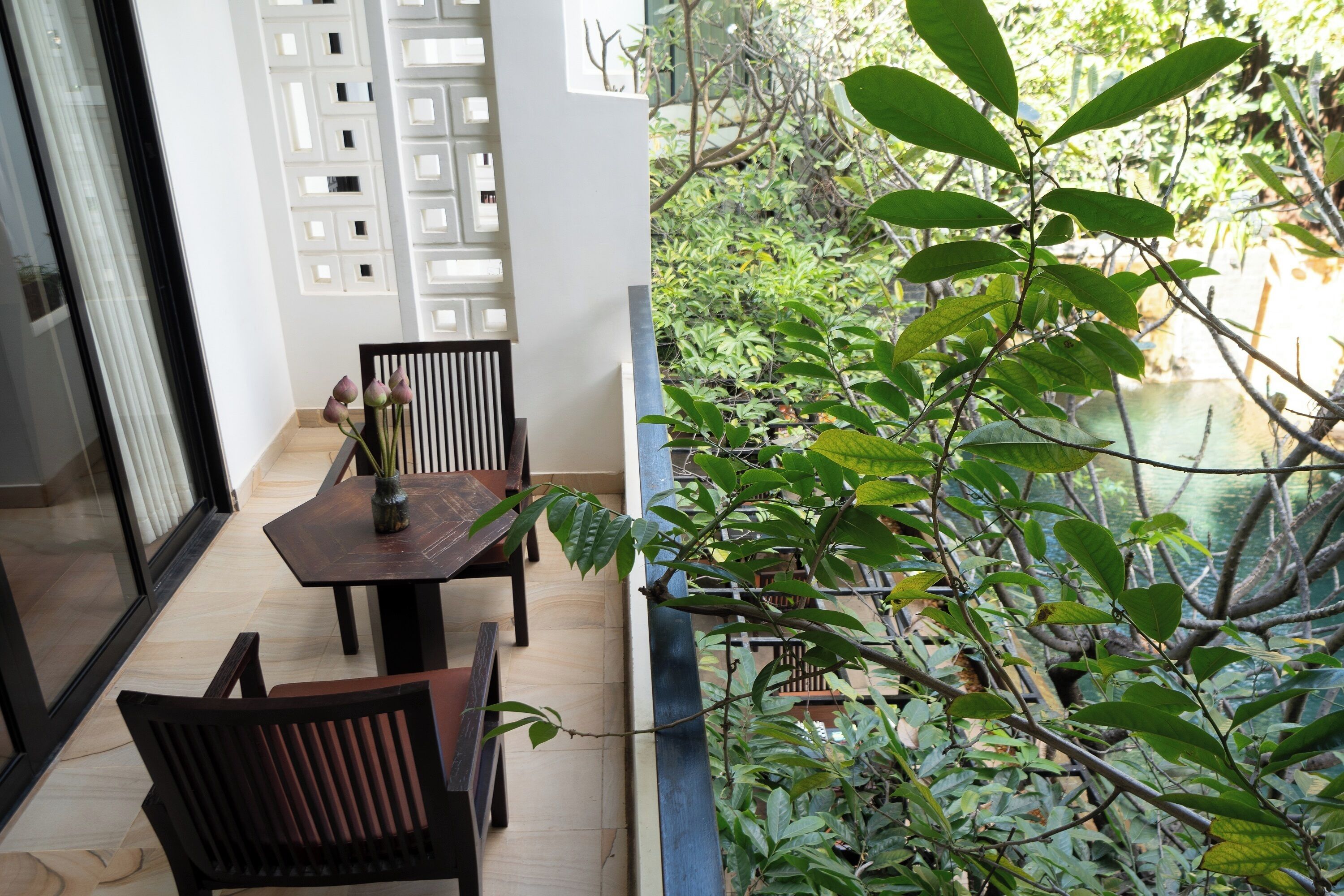 A balcony with a view of a water body and greenery, a dining area with a table and chairs, and a white partition with a geometric design.