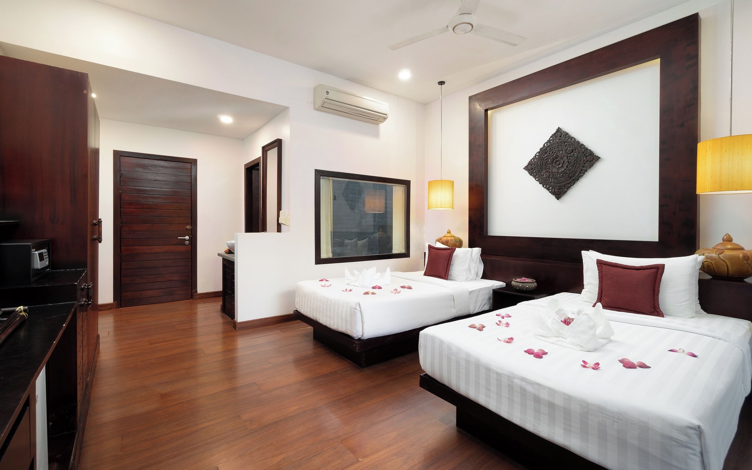 A hotel room with two beds, wooden flooring, a ceiling fan, an air conditioning unit, a large window, and a decorative wall piece.
