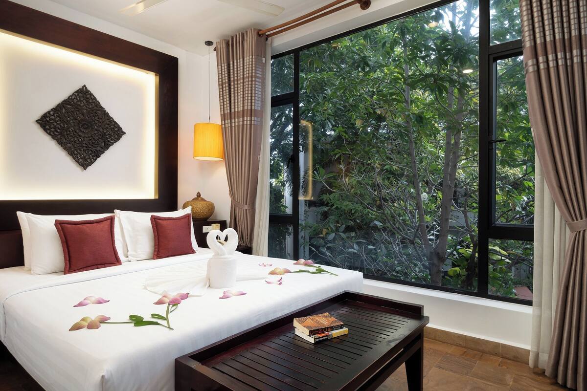 Quarto de hotel com uma cama grande, uma mesa de cabeceira, uma luminária e vista para o verde através de uma janela ampla.
