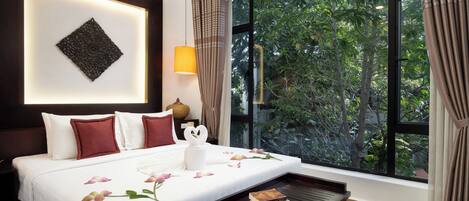 Une chambre d’hôtel avec un grand lit, une table de chevet, une lampe et une vue sur la verdure grâce à une grande fenêtre.