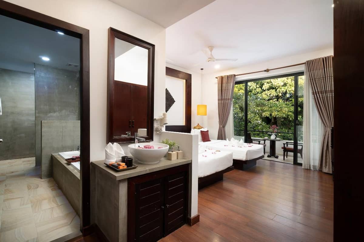 Quarto de hotel com um espelho grande, pia, cama, mesa pequena e varanda com vista para o verde.