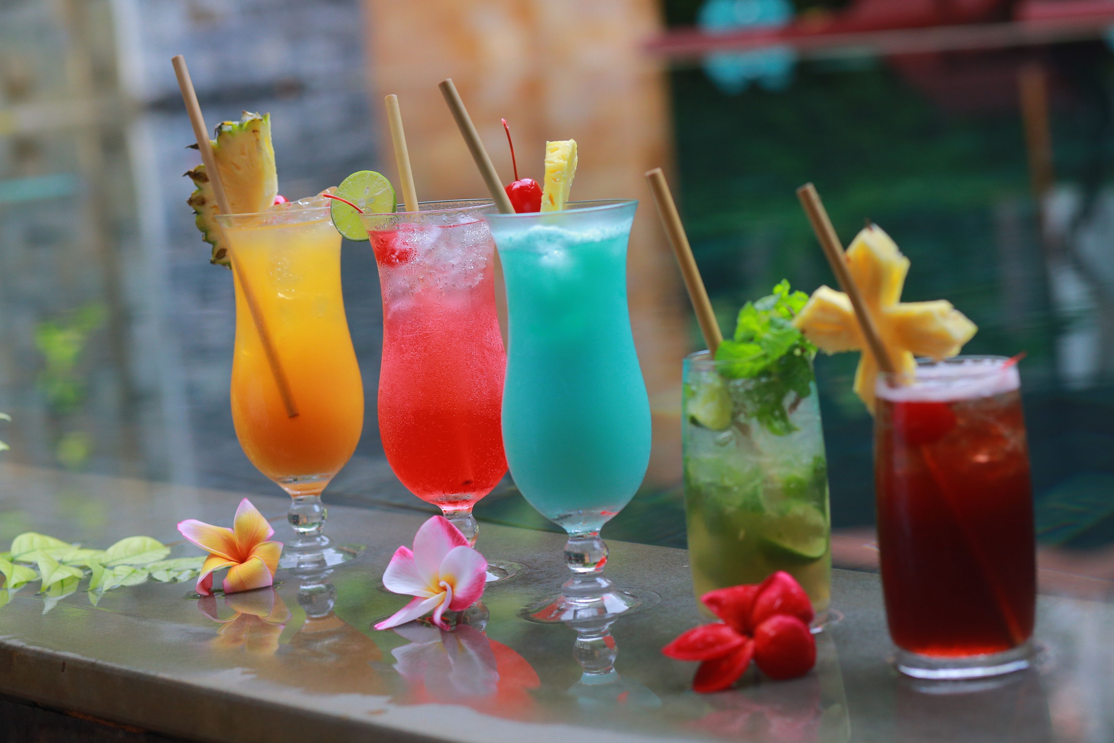 Quatre cocktails colorés, servis avec des pailles et des décorations, sont présentés sur une surface réfléchissante.
