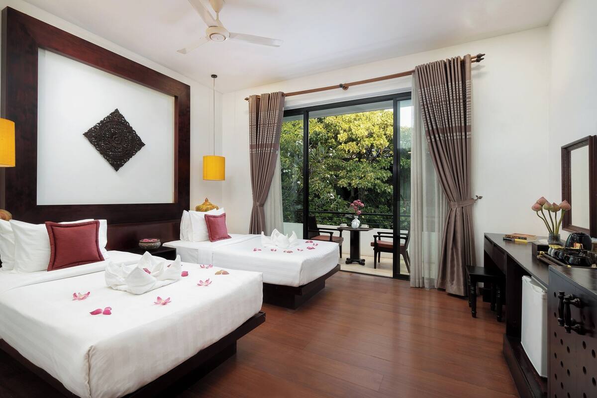 Quarto de hotel com duas camas, uma escrivaninha, uma cadeira, uma mesinha e uma varanda com cortinas.