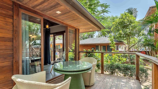A wooden deck with wicker furniture and a glass table, overlooking a lush garden. - The Baga Beach Resort (Baga)