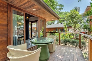 Terraza de madera con muebles de mimbre y una mesa de vidrio, con vistas a un jardín exuberante.