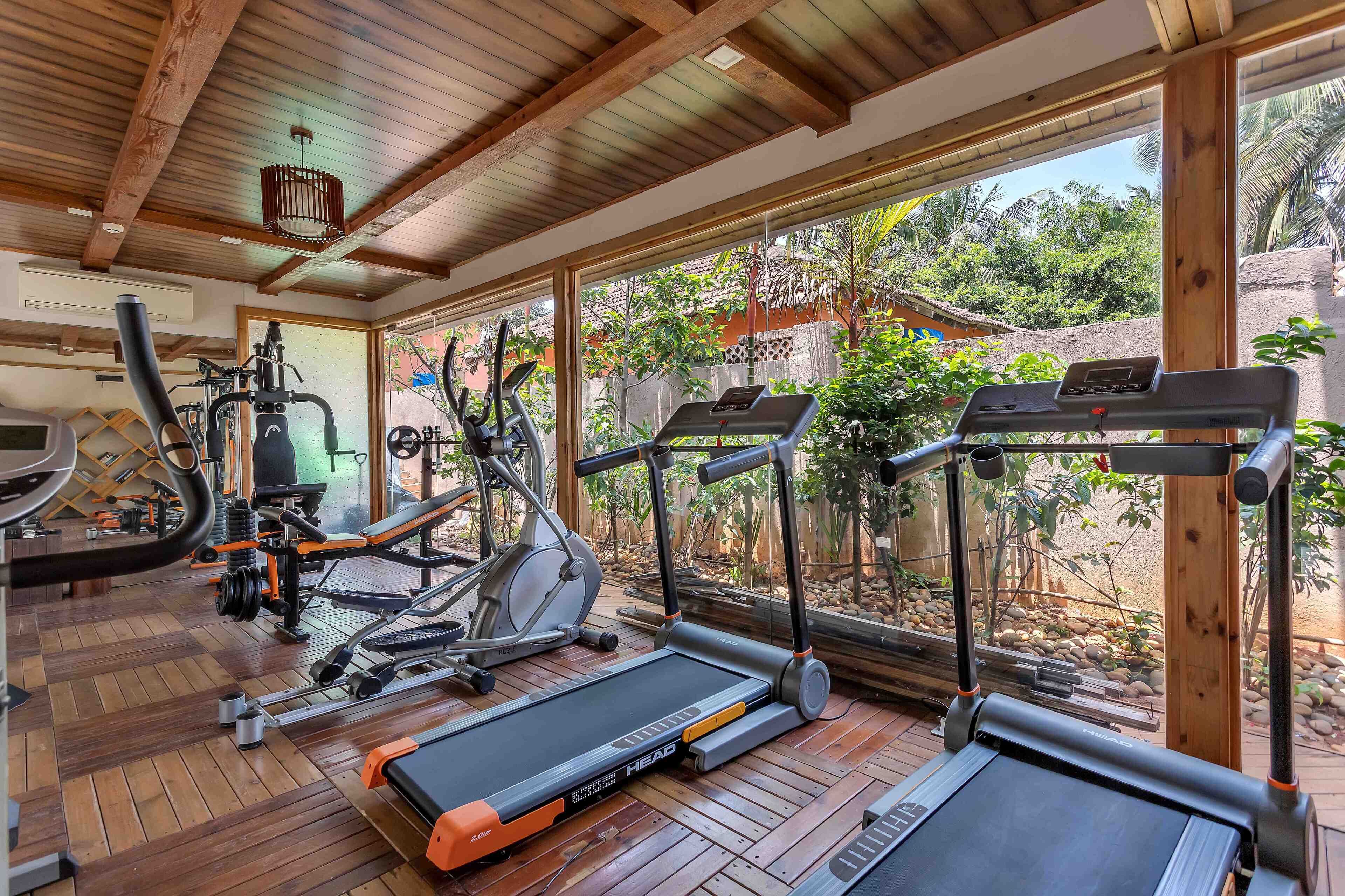 A gym with treadmills, an elliptical, and weight machines, featuring large windows and wooden floors.