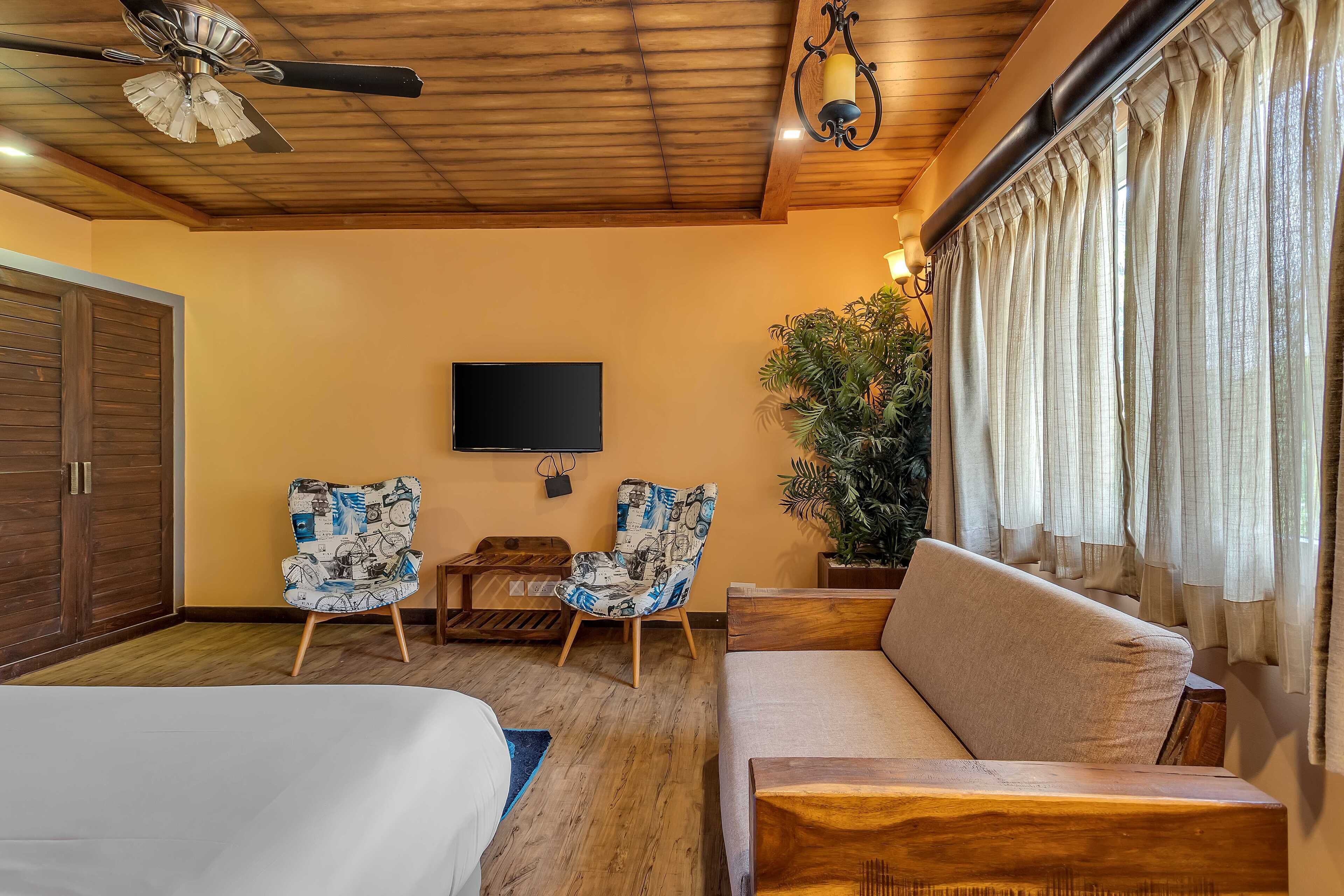 A room with a wooden ceiling, a ceiling fan, a TV mounted on the wall, two patterned chairs, a sofa, a potted plant, and a window with curtains.