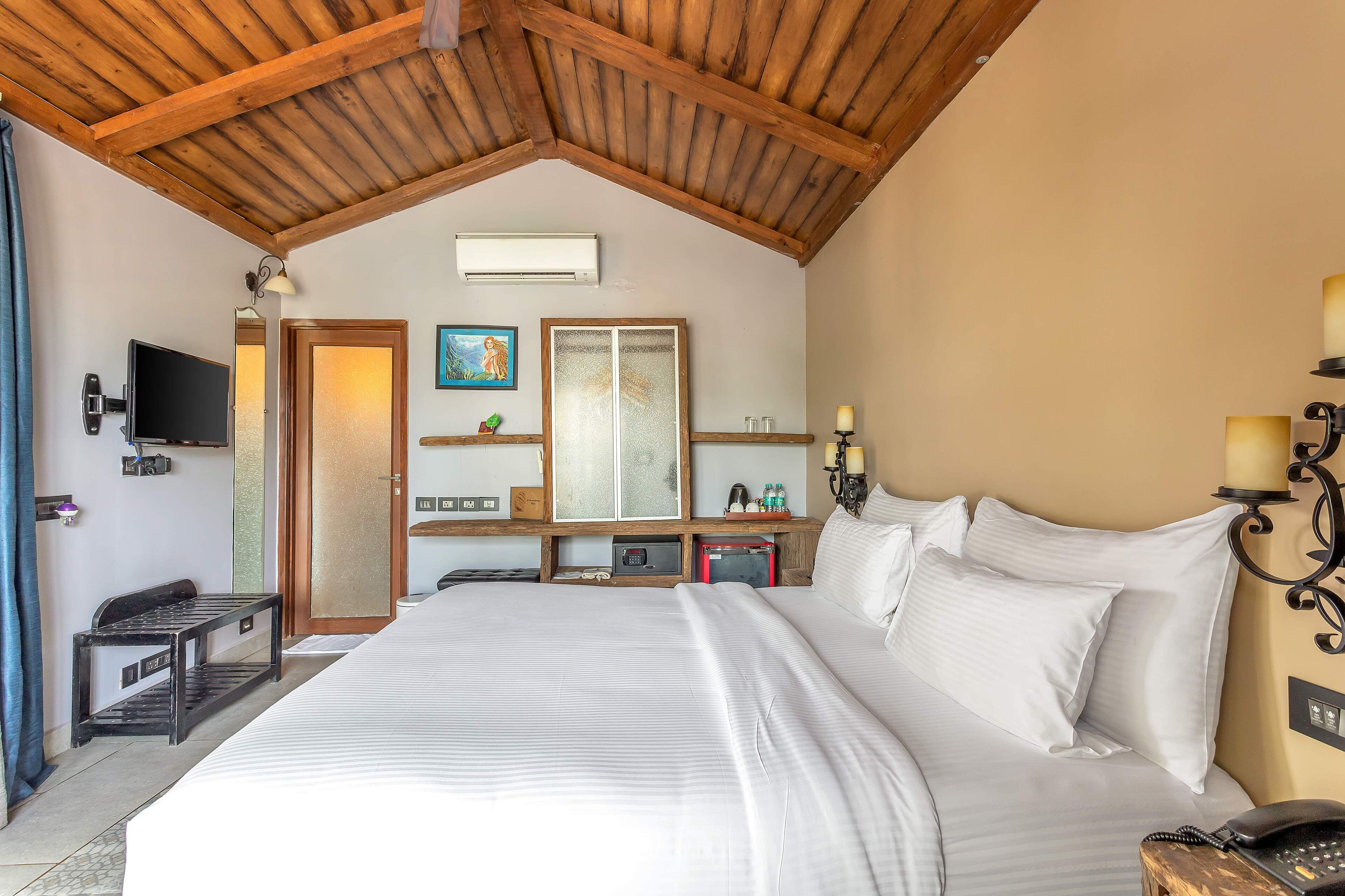 A bedroom with a large bed, wooden ceiling beams, a TV, bedside lamps, a desk with a computer, and a mirror.