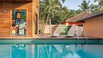 Un área de piscina con sillones reclinables, un edificio de madera con obras de arte y palmeras al fondo.