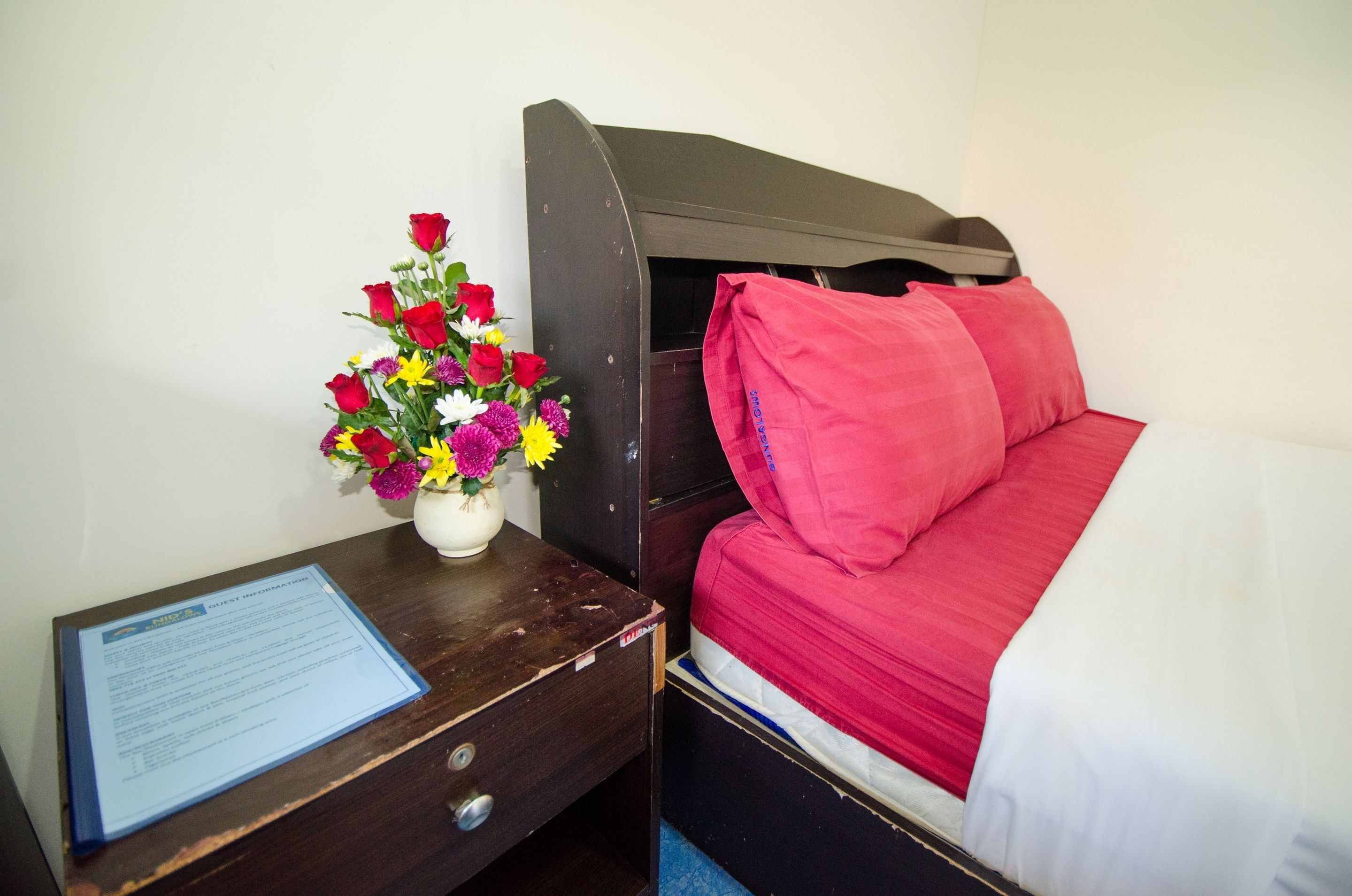 Habitación de hotel pequeña y funcional con una cama individual, una mesita de noche con un florero y un escritorio con un documento.