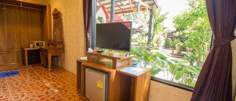 Habitación con suelo de baldosas, un escritorio de madera con televisor, un refrigerador pequeño y ventana con vistas a un jardín.