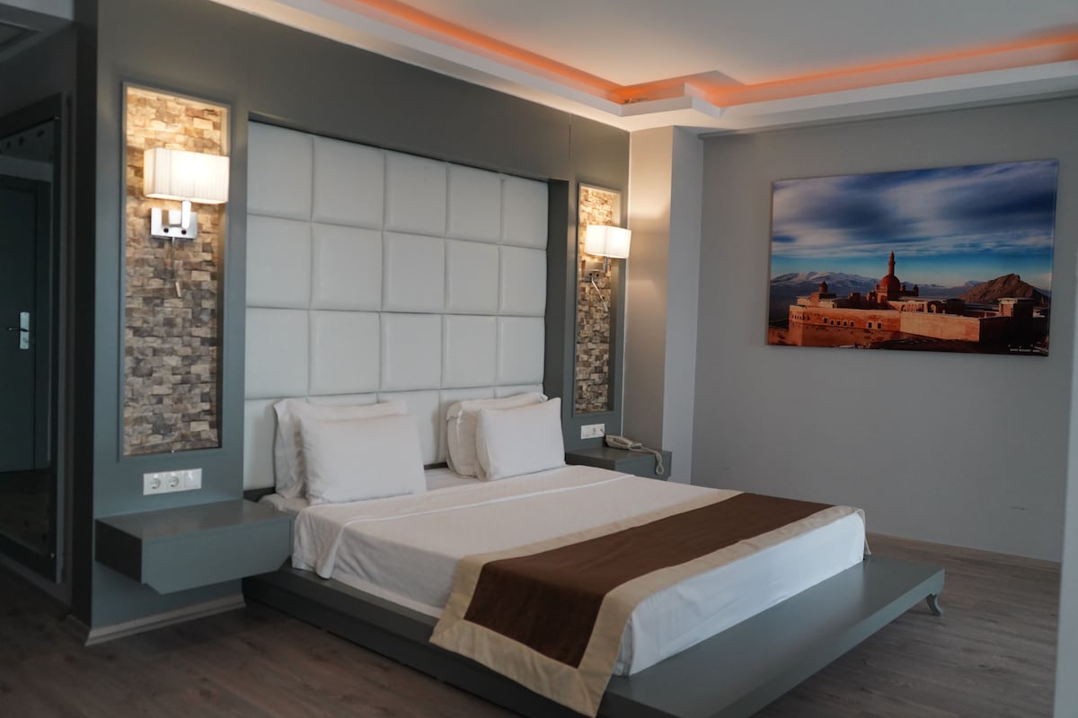 Una habitación de hotel moderna con una cama grande, cabecero integrado, lámparas de noche, mesitas de noche y un cuadro colgado en la pared.