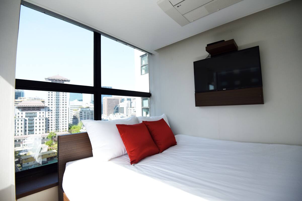 Una habitación de hotel con un ventanal grande, una cama con almohadas blancas y rojas, y un televisor de pantalla plana montado en la pared.