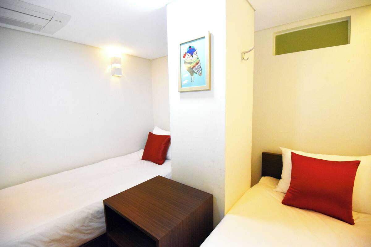 Habitación de hotel con dos camas individuales, una mesita de noche de madera, una lámpara de pared y un cuadro enmarcado.