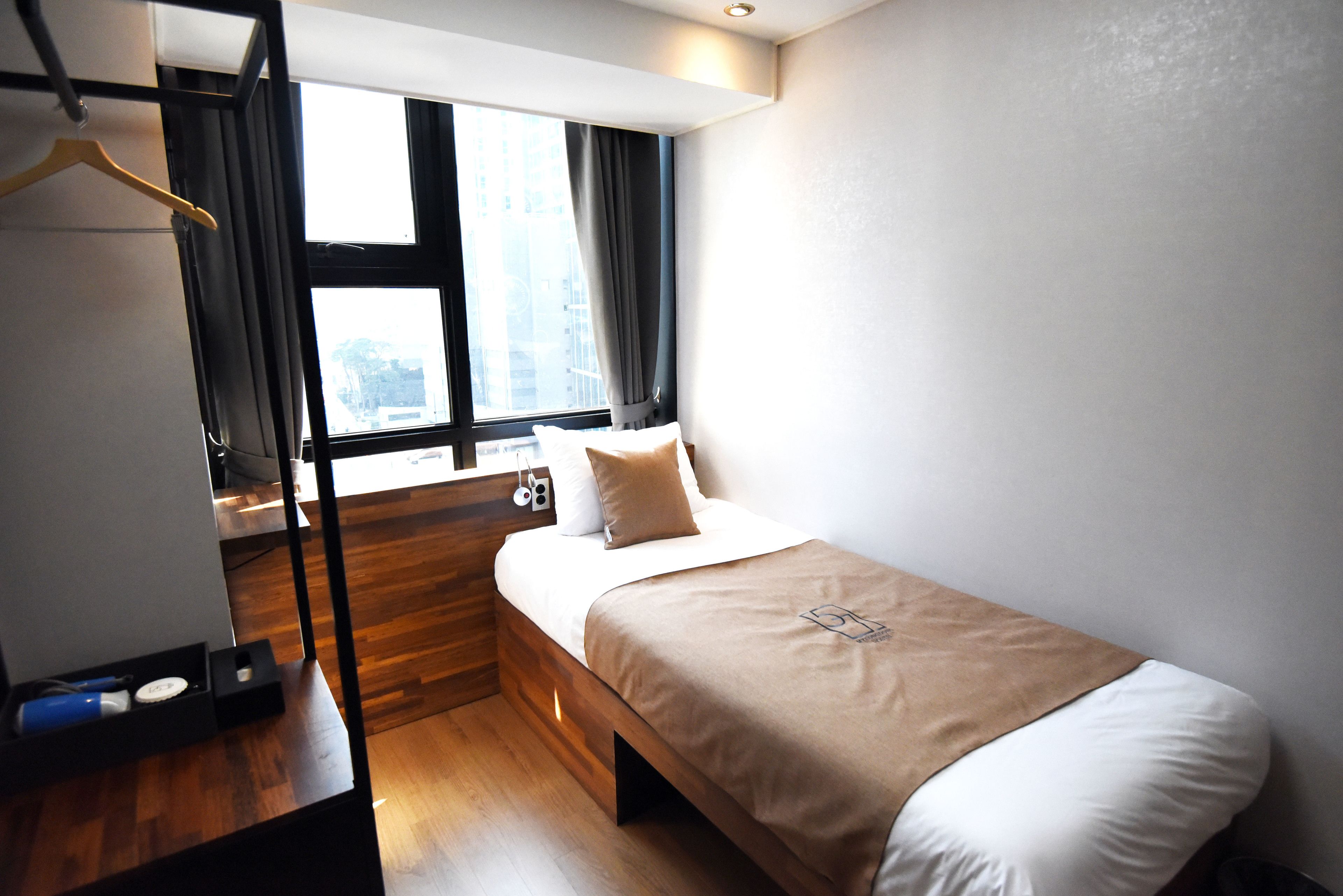 A compact hotel room with a single bed, a wooden nightstand, a desk with a chair, a window with curtains, and a coat rack.