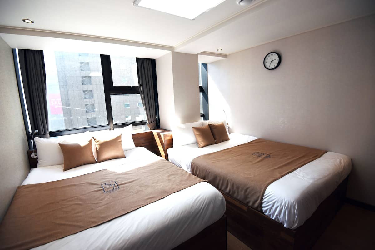 Habitación de hotel con dos camas, una ventana con cortinas, un reloj en la pared y un tragaluz.