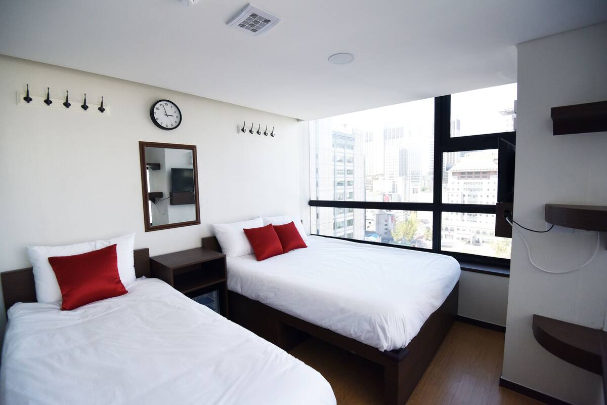 Habitación de hotel con dos camas, un ventanal con vistas a la ciudad, un reloj y un espejo en la pared.