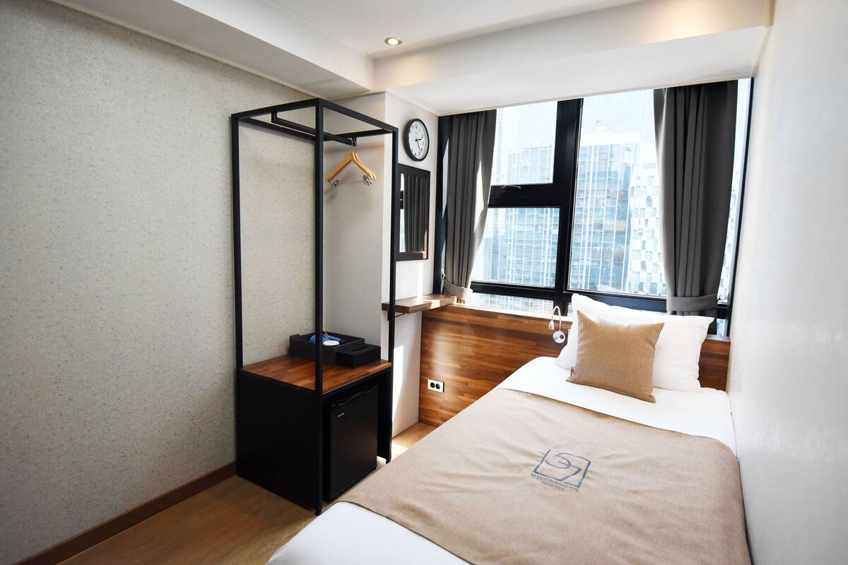 Habitación de hotel moderna con una cama, un escritorio de madera, un perchero y una ventana con vistas a la ciudad. También se observa un reloj en la pared.