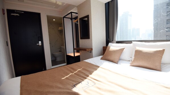 Habitación de hotel con cama, ventana con vista a la ciudad, baño con inodoro y ducha, y una puerta.