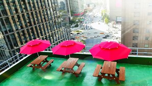 Un balcone con tavoli da picnic in legno e ombrelloni rosa, con vista su una strada trafficata e palazzi alti.