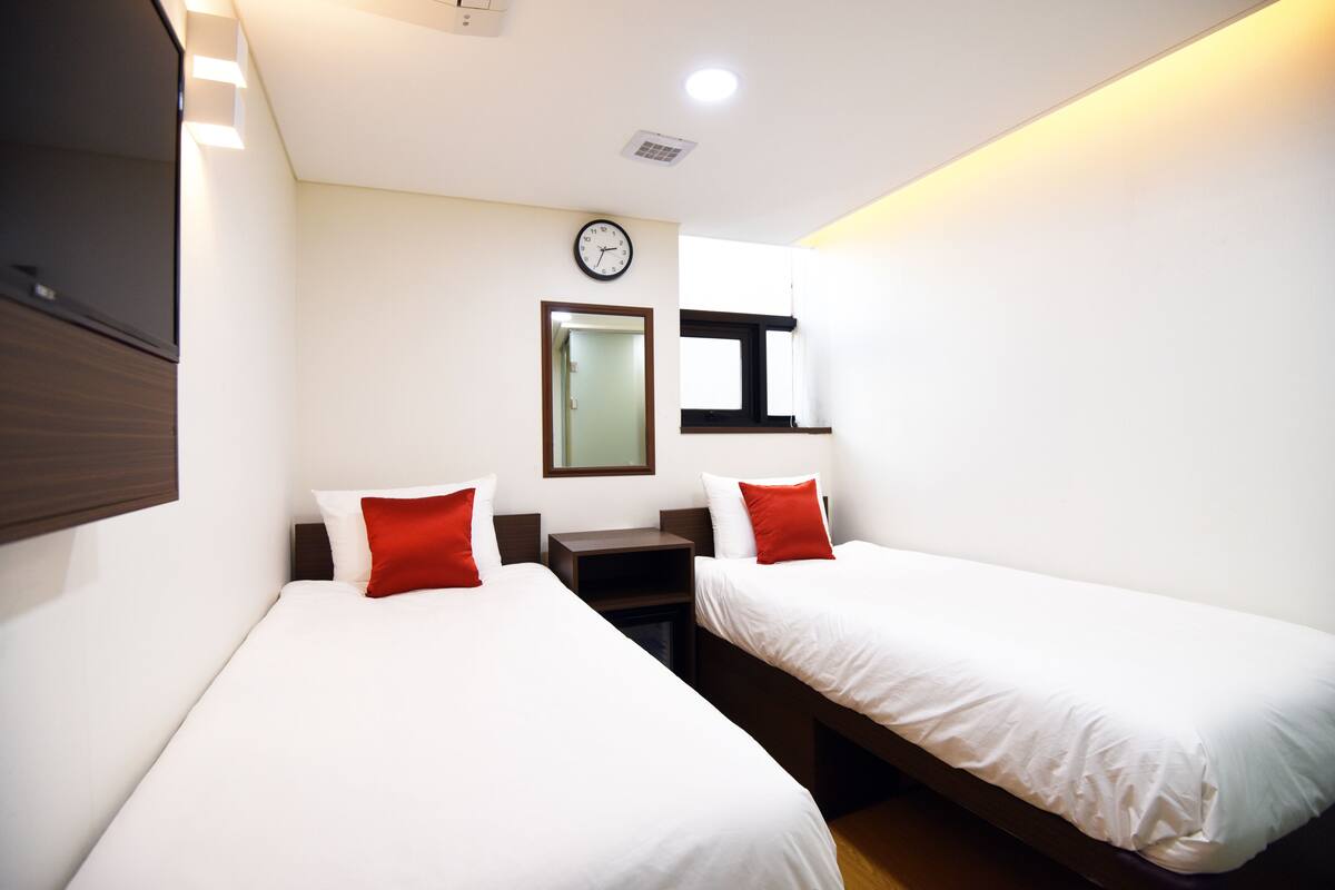Una habitación de hotel con dos camas individuales, una mesita de noche, un reloj en la pared y una ventana con persianas.