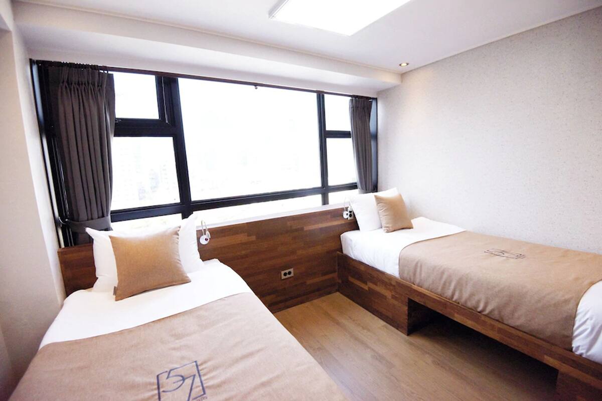 Habitación de hotel con dos camas individuales, un ventanal grande con cortinas, cabecero de madera y pared de color claro.