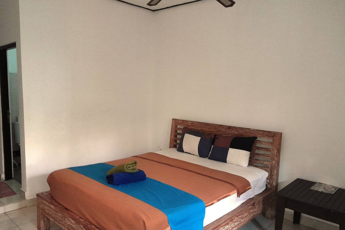 Un lit simple avec une tête de lit en bois, une couvre-lit bleue et orange, et une petite table de chevet sur laquelle se trouve un magazine.