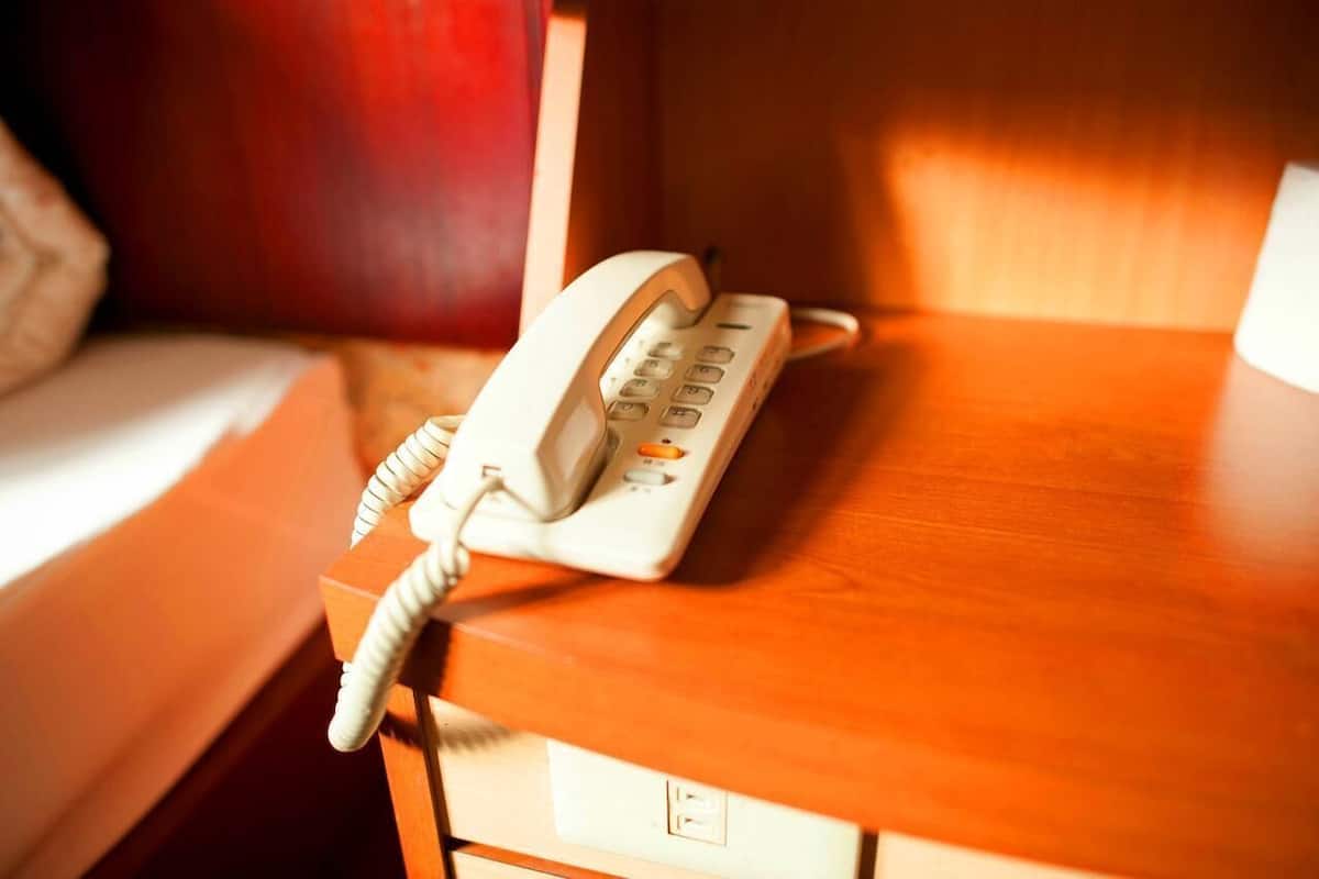 A hotel room with a wooden bedside table, a white telephone, and a bed with a white headboard.