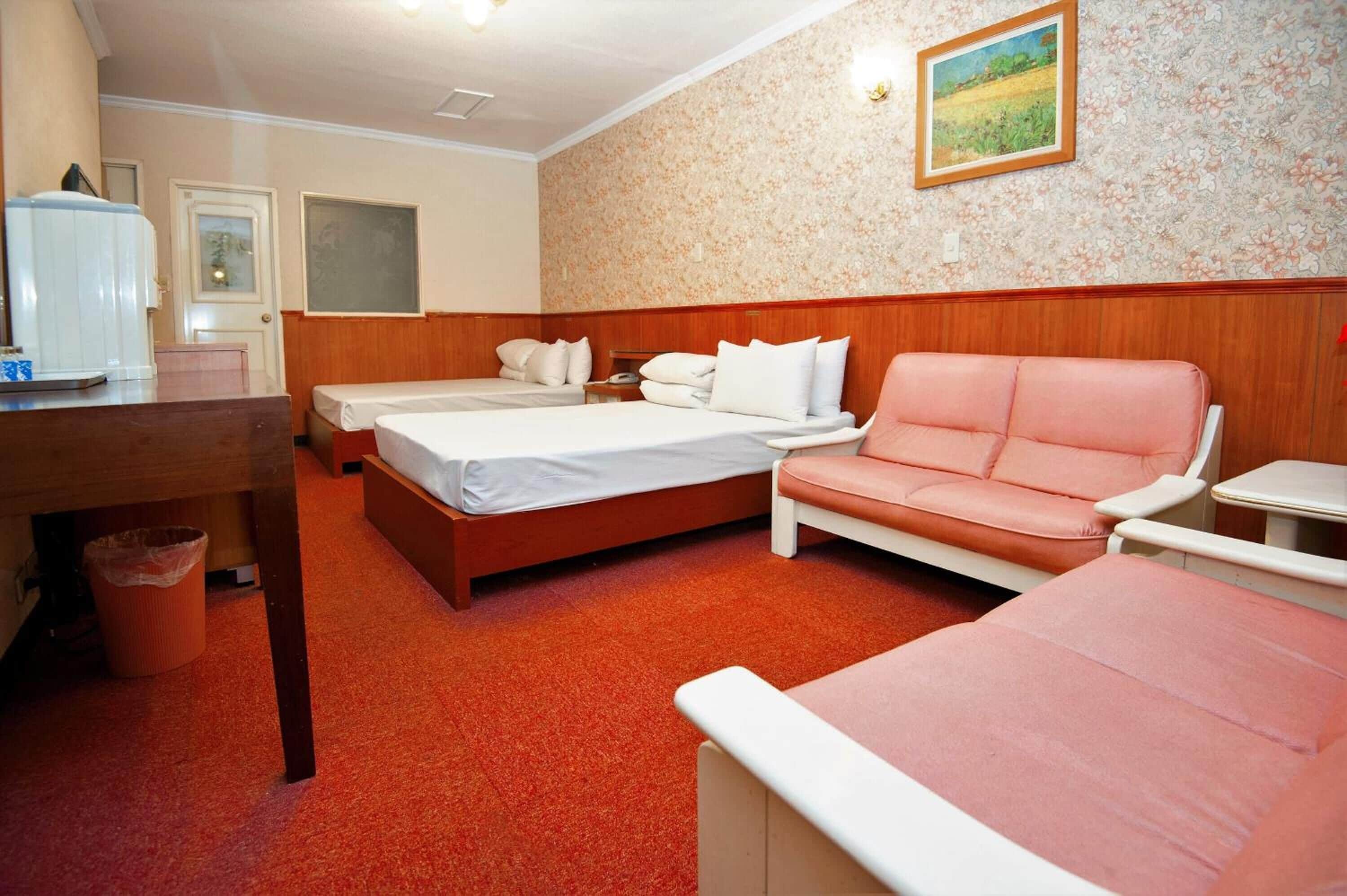 A hotel room with two beds, a sofa, a small table, and a trash bin.