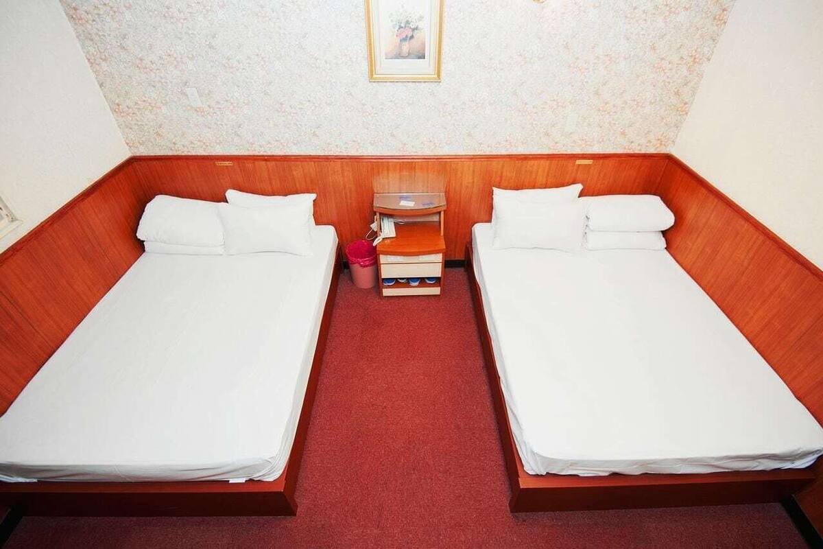 A room with two beds, a wooden headboard, a small table with a red bucket, and a framed picture on the wall.